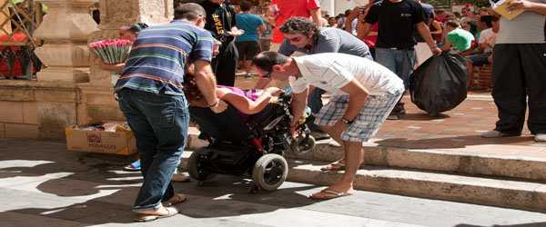 Una persona usuaria de silla de ruedas es ayudada por varias personas ante la falta de accesibilidad al recinto que quiere entrar
