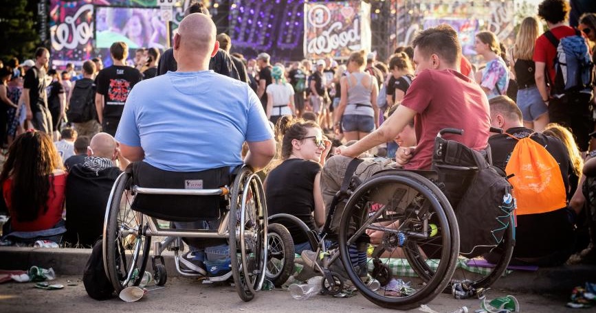 Personas en silla de ruedas disfrutando de un festival entre el público, con escenario y ambiente festivo de fondo.