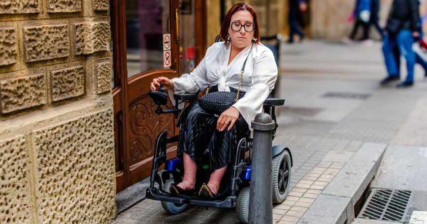 Mujer en silla de ruedas eléctrica frente a un bolardo y un escalón que impiden su paso en una calle de Madrid.