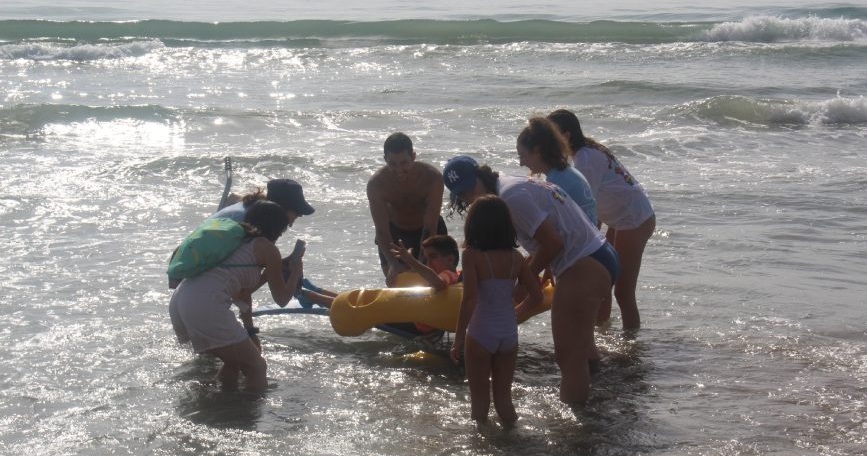 Grupo de voluntariado acompaña a una persona con discapacidad en silla anfibia para disfrutar del mar.