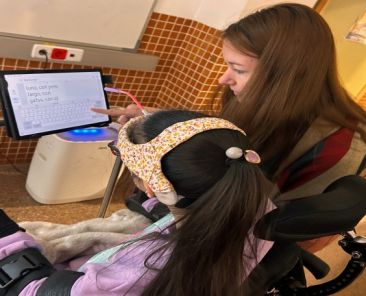 Apoyo educativo inclusivo con tecnología accesible Educadora apoya a alumna con discapacidad mientras usa una tableta adaptada para comunicarse.