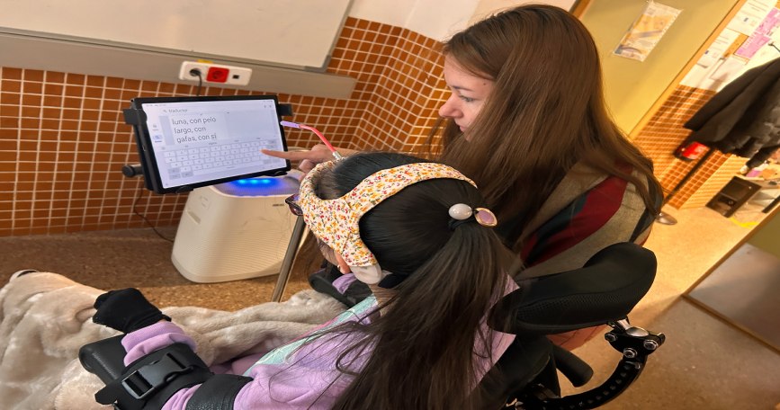 Educadora apoya a alumna con discapacidad mientras usa una tableta adaptada para comunicarse.
