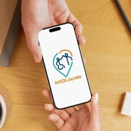 Persona sostiene un móvil con el logotipo de la app InVOLúcrate by Cocemfers sobre una mesa de trabajo.