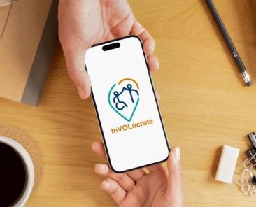 Persona sostiene un móvil con el logotipo de la app InVOLúcrate by Cocemfers sobre una mesa de trabajo.