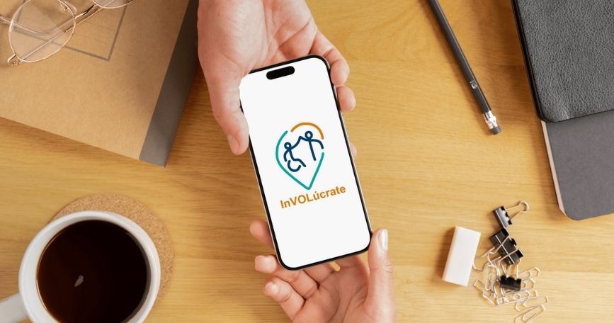 Persona sostiene un móvil con el logotipo de la app InVOLúcrate by Cocemfers sobre una mesa de trabajo.