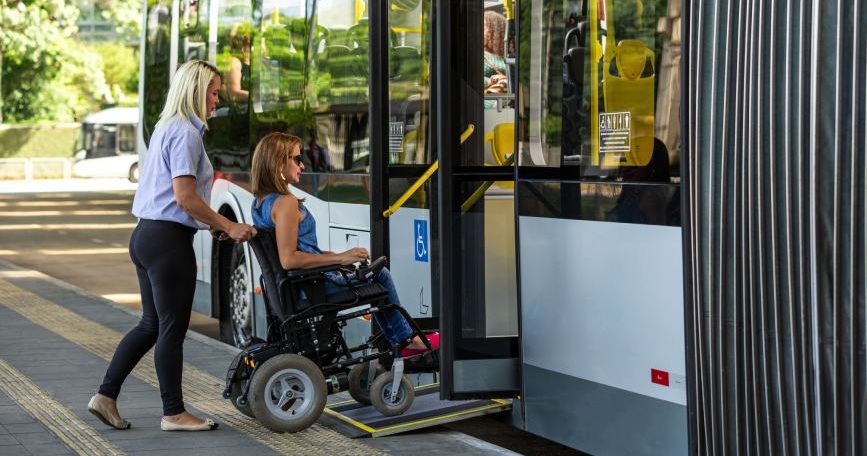 Mujer en silla de ruedas entrando a un autobús mediante una rampa asistida por una trabajadora.