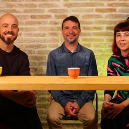 Grabación del podcast “Voces con FQ” Tres personas participan en la grabación del podcast “Voces con FQ”, sentadas ante una mesa con micrófonos y tazas de colores en un estudio iluminado.