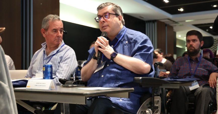 Javier Font, sentado en su silla de ruedas, toma la palabra con un micrófono durante un acto en una sala de conferencias.