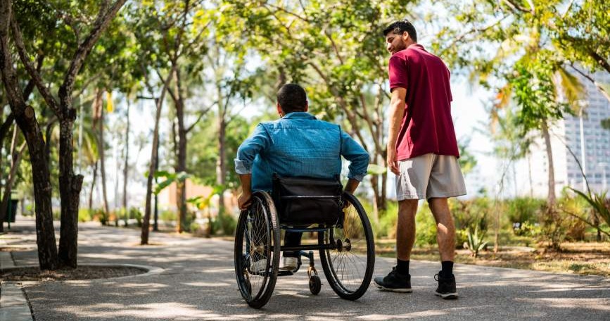 Persona usuaria de silla de ruedas paseando por un parque junto a otra persona que la acompaña.
