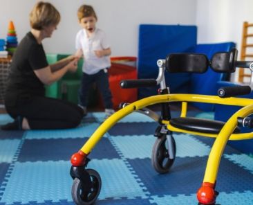 Niño con discapacidad motora realizando una actividad de atención temprana junto a una profesional, con un andador adaptado en primer plano.