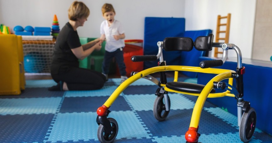 Niño con discapacidad motora realizando una actividad de atención temprana junto a una profesional, con un andador adaptado en primer plano.