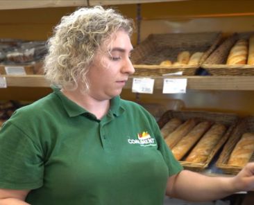 Persona trabajadora usando un terminal de venta en una panadería con estanterías de pan al fondo.