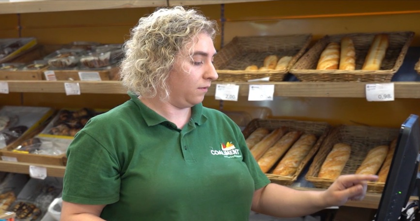 Persona trabajadora usando un terminal de venta en una panadería con estanterías de pan al fondo.