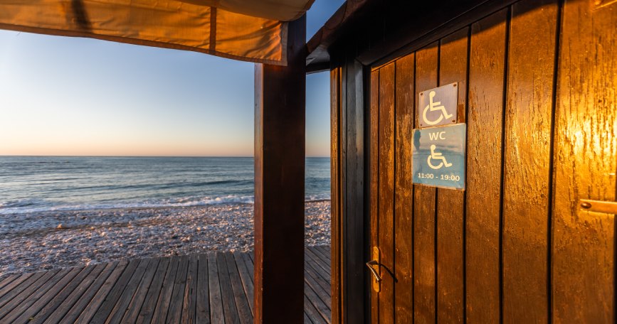 Puerta de aseo accesible en una playa con señal de discapacidad y el mar al fondo al atardecer