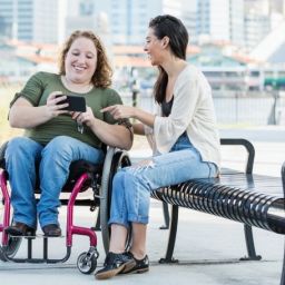 Persona usuaria de silla de ruedas utilizando un móvil junto a otra persona en un entorno urbano accesible.