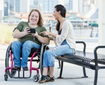 Persona usuaria de silla de ruedas utilizando un móvil junto a otra persona en un entorno urbano accesible.