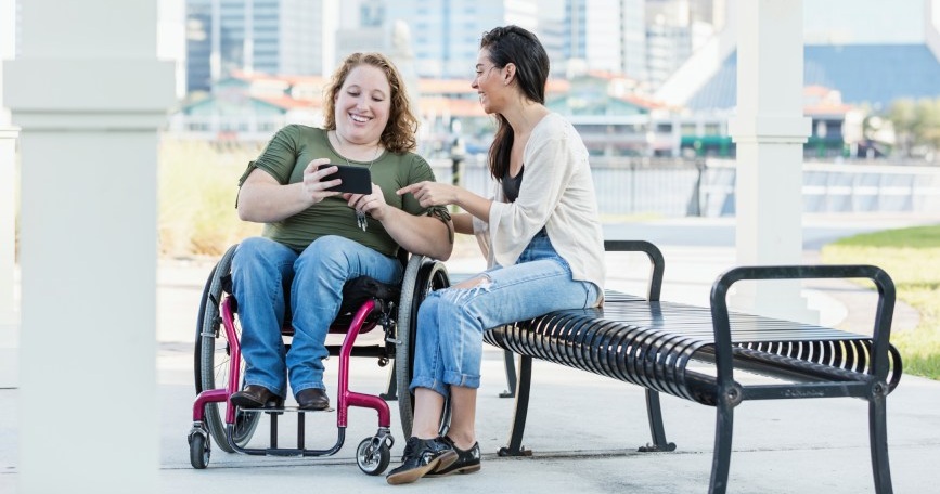 Persona usuaria de silla de ruedas utilizando un móvil junto a otra persona en un entorno urbano accesible.