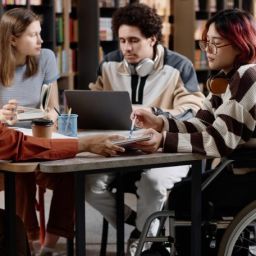 Grupo diverso de personas jóvenes, incluida una mujer usuaria de silla de ruedas, colaborando en una mesa de trabajo en un entorno educativo inclusivo.