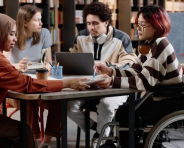 Grupo diverso de personas jóvenes, incluida una mujer usuaria de silla de ruedas, colaborando en una mesa de trabajo en un entorno educativo inclusivo.