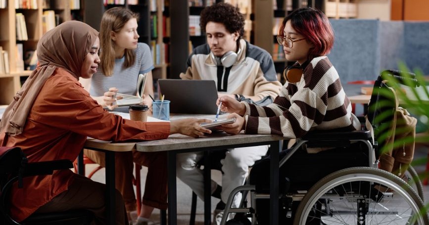 Grupo diverso de personas jóvenes, incluida una mujer usuaria de silla de ruedas, colaborando en una mesa de trabajo en un entorno educativo inclusivo.