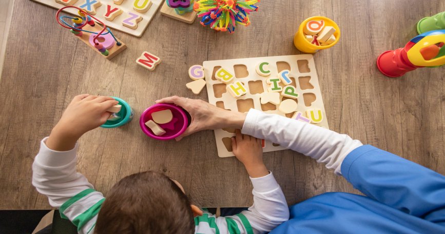 Menor realizando una actividad de aprendizaje con letras y juguetes educativos junto a una profesional de atención temprana.