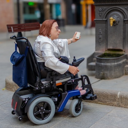 Persona usuaria de silla de ruedas intentando acceder a una fuente pública en una calle, ejemplo de barreras de accesibilidad en el entorno urbano.