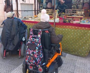 Dos personas en silla de ruedas interactúan en un puesto de mercado en Deusto, fomentando la inclusión comunitaria
