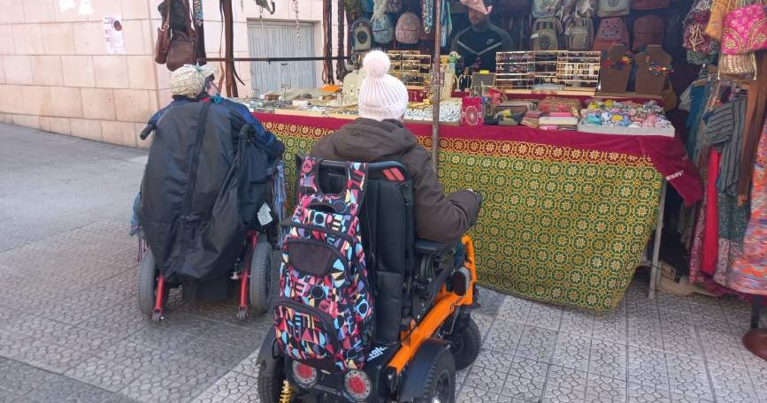 Dos personas en silla de ruedas interactúan en un puesto de mercado en Deusto, fomentando la inclusión comunitaria
