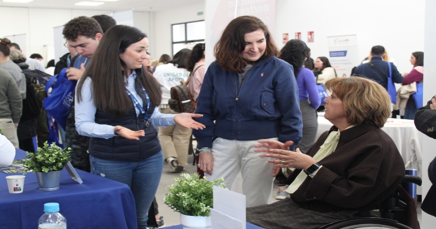 Participantes conversan en un estand de la IV Feria de Empleo de COCEMFE Sevilla con una mujer usuaria de silla de ruedas.