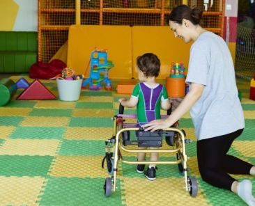 Atención Temprana a menor con discapacidad en entorno adaptado Profesional apoya a un niño con discapacidad en ejercicios de Atención Temprana con andador en sala adaptada