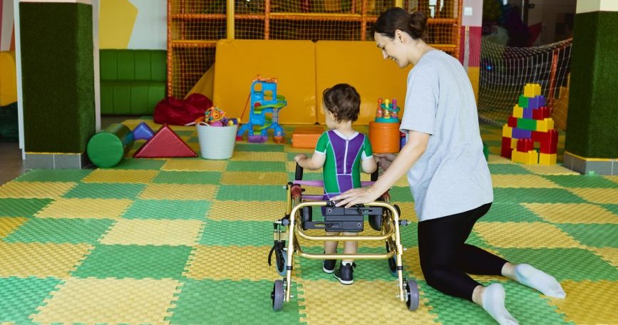 Profesional apoya a un niño con discapacidad en ejercicios de Atención Temprana con andador en sala adaptada