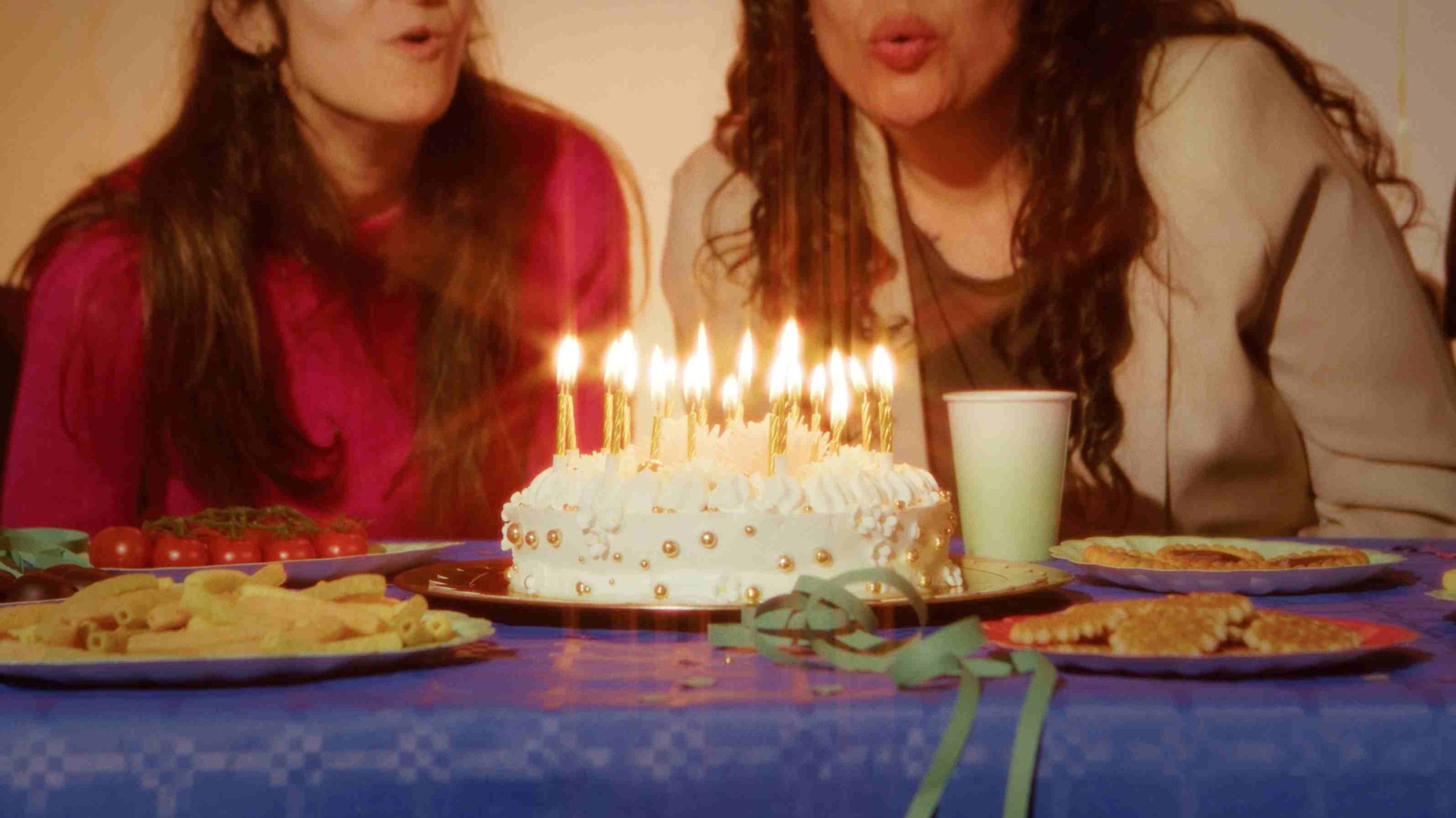 Dos mujeres soplan velas de una tarta en una celebración con comida sobre la mesa