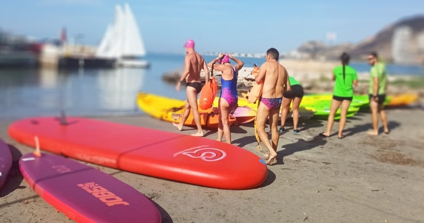Personas preparándose para deporte adaptado con tablas y kayaks en la playa