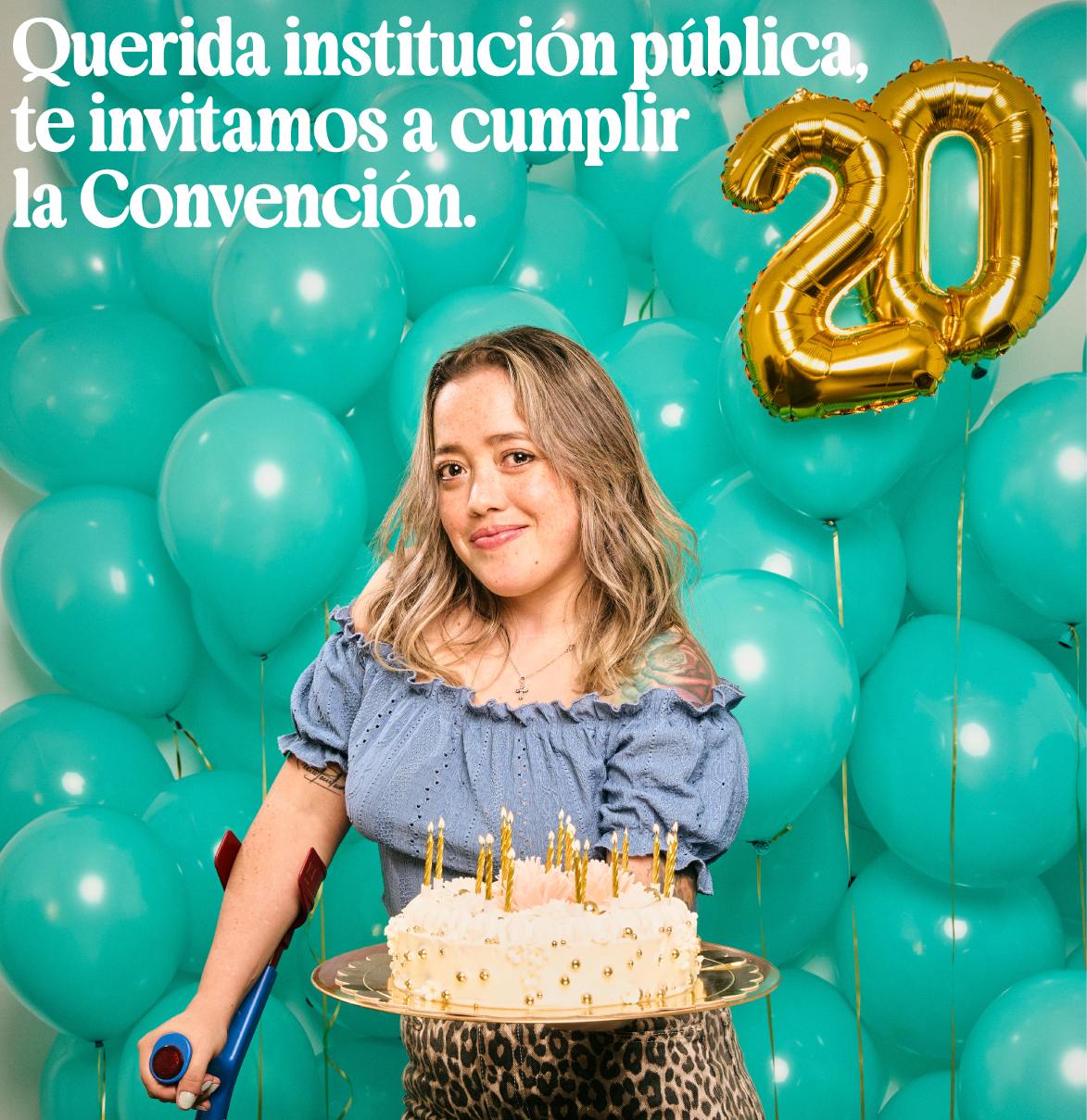 Mujer con acondroplasia sostiene una tarta y una muleta ante globos del 20 aniversario