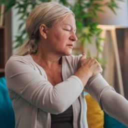 Mujer con dolor muscular relacionado con fibromialgia Mujer sentada en un sofá sujetándose el hombro debido al dolor muscular asociado a la fibromialgia.