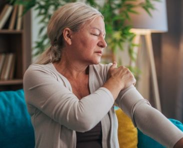 Mujer sentada en un sofá sujetándose el hombro debido al dolor muscular asociado a la fibromialgia.