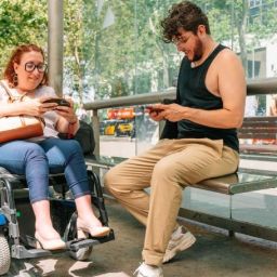 Mujer usuaria de silla de ruedas eléctrica y otra persona en una parada accesible utilizando teléfonos móviles.