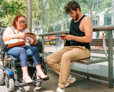 Asistencia personal, accesibilidad y vida independiente Mujer usuaria de silla de ruedas eléctrica y otra persona en una parada accesible utilizando teléfonos móviles.