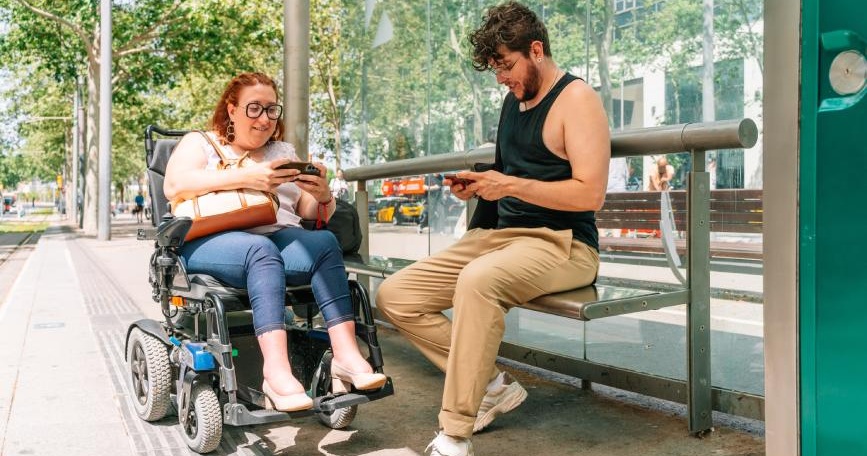 Mujer usuaria de silla de ruedas eléctrica y otra persona en una parada accesible utilizando teléfonos móviles.