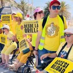Personas con discapacidad reivindican playas accesibles en Alicante con carteles amarillos en la playa del Postiguet.