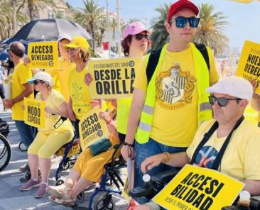 Personas con discapacidad reivindican playas accesibles en Alicante con carteles amarillos en la playa del Postiguet.