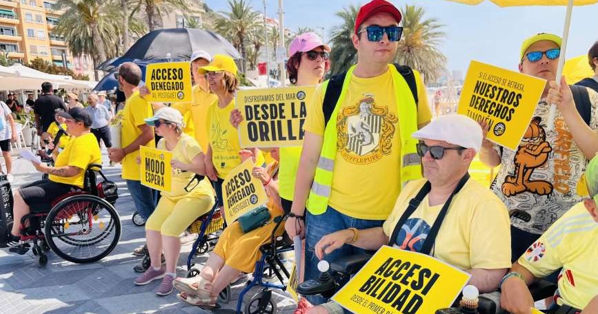 Personas con discapacidad reivindican playas accesibles en Alicante con carteles amarillos en la playa del Postiguet.