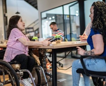 Inclusión laboral de las personas con acondroplasia Mujer con acondroplasia, usuaria de silla de ruedas, participa en una reunión de trabajo en una oficina accesible.