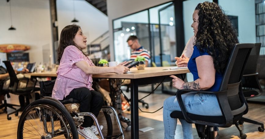 Mujer con acondroplasia, usuaria de silla de ruedas, participa en una reunión de trabajo en una oficina accesible.