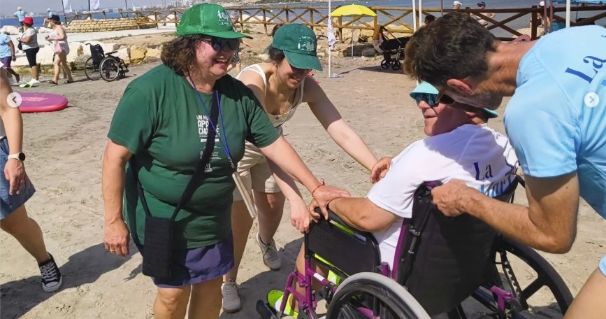 Persona usuaria de silla de ruedas participa en una actividad en la playa junto a personas voluntarias durante La Mar Solidaria en Alicante.