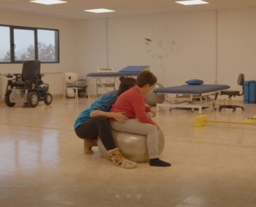 Profesional realiza una sesión de fisioterapia con un niño con discapacidad en una sala de rehabilitación equipada con material terapéutico y ayudas técnicas.