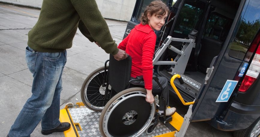 Persona usuaria de silla de ruedas accediendo a un vehículo adaptado mediante una plataforma elevadora con apoyo de otra persona.