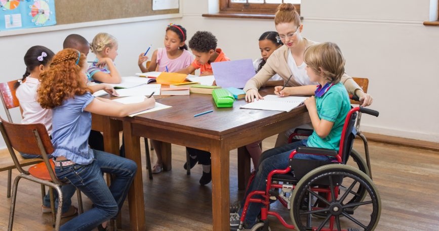 Docente y grupo de escolares, entre ellos un alumno usuario de silla de ruedas, participan juntos en una actividad en un aula inclusiva.