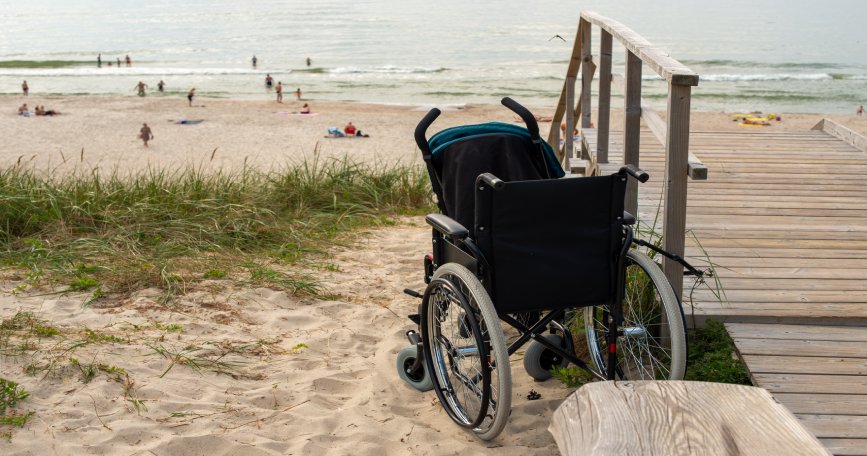 Silla de ruedas junto a una pasarela de acceso a una playa con el mar al fondo, representando la necesidad de mejorar la accesibilidad del litoral.