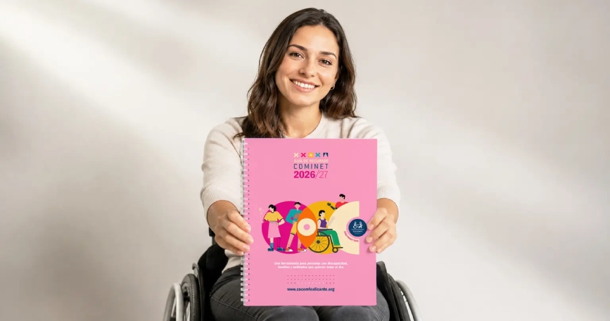 Una mujer usuaria de silla de ruedas sostiene la Guía de Recursos COMINET 2026/27 de COCEMFE Alicante, una publicación gratuita con información sobre recursos, servicios y prestaciones de la provincia de Alicante.